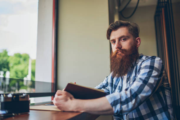 istock Half length portrait of skilled bearded student with textbook in hand looking at camera while sitting in coffee shop and preparing for upcoming exams.Publicity area for your advertising text message 1159408322