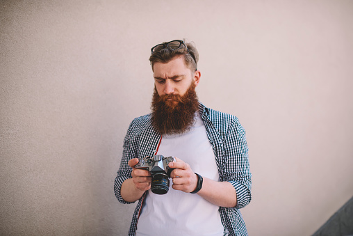 Bearded stylish amateur with eyewear on head making settings on vintage camera and browsing photos standing in urban setting against promotional background.Photographer checking light on device