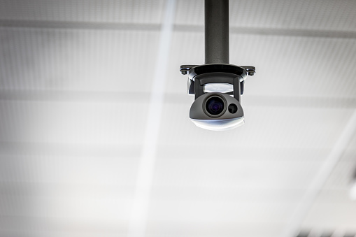 Overhead surveillance camera suspended from the ceiling hanging on a bracket in a grey monochrome image with vignette and copy space