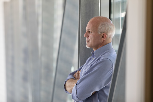 Senior grey-haired man standing deep in thought in front of a window with folded arms and a deadpan serious expression, close up in profile indoors