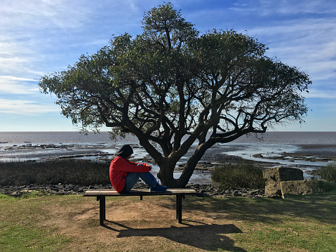 Lonely sad man sitting on park bench next to the Rio de la Plata river in Buenos Aires province