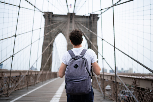 This is a color photograph of a young American man wearing a backpack as he walks across the Brooklyn Bridge in New York City, USA.