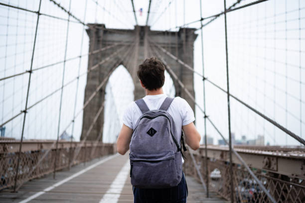 Millennial Man with Backpack Walking Across the Brooklyn Bridge stock photo