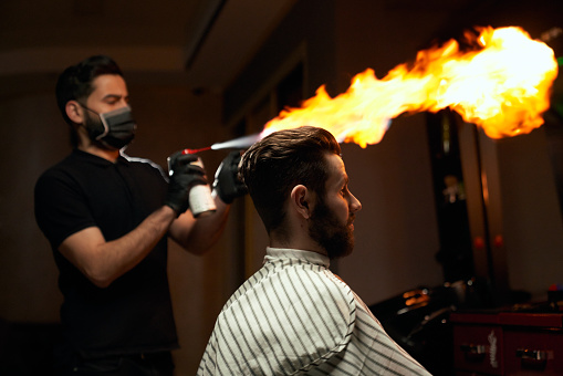 Portrait of stylish male bearded client sitting in chair and waiting while proficient hairdresser is making all preparation for haircut with fire. Concept of extreme masculinity service in barbershop
