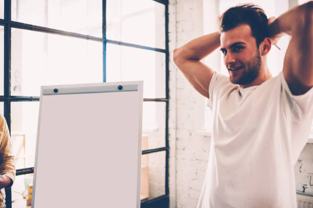 istock Handsome male student standing near blank board preparing for explaining his ideas for startup, happy hipster guy thinking over making speech and using flip chart for presentation creative solutions 1159423478