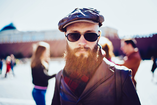 Close up portrait of bearded hipster guy in sunglasses dressed in trendy apparel looking at camera during strolling on city streets, stylish tourist spending vacations in Russia enjoying weekend