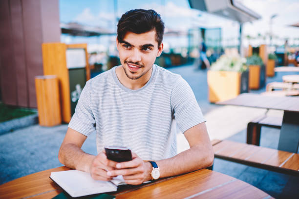 istock Pondering cheerful blogger thinking on funny publication on internet website holding smartphone.Casual dressed student looking away while chatting online on cellular sitting at wooden table with book 1159431069