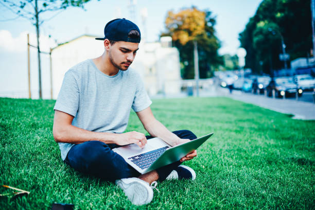 istock Pensive student in casual outfit sitting on green grass with laptop computer and checking mail via free 4G internet on university territory.Male blogger in trendy cap updating software on netbook 1159431146