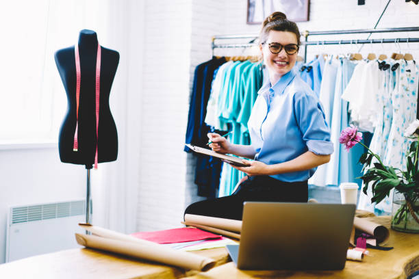 istock Smiling female designer in eyeglasses looking at camera while making notes of measurements working in showroom on collection of stylish clothing sitting on desktop with garment accessories and netbook 1159432756