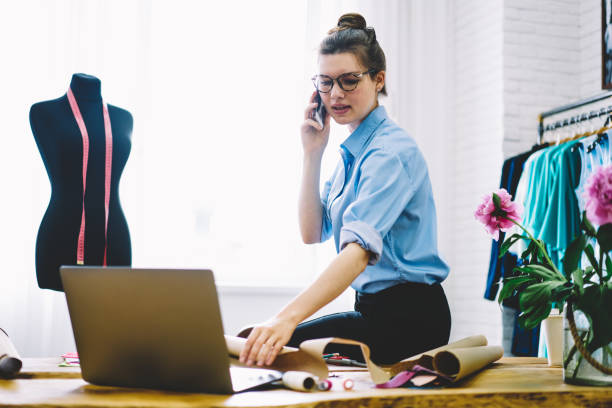 istock Manager organizing working schedule of showroom on telephone sitting on sewing table.Creative dressmaker talking on smartphone with designer to preparing collection for fashion industry event 1159432812