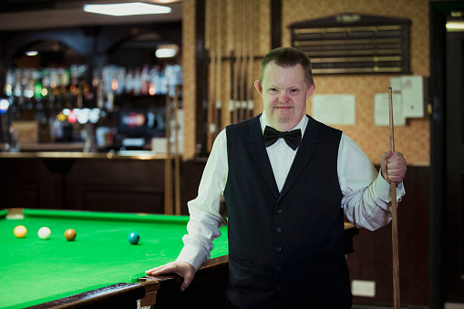 Well dressed Man with Down syndrome holding a pool cue, leaning on a pool table.