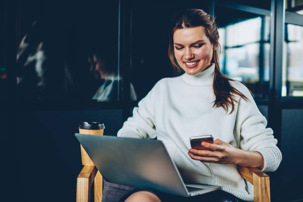 istock Cheerful female blogger sharing information via smartphone and laptop devices via bluetooth connection,smiling successful hipster girl reading positive email message from friend resting at coffee shop 1159433173