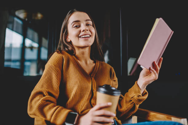 Portrait of successful smiling woman looking at camera and laughing holding caffeine beverage and notebook for knowledge, positive cheerful hipster girl enjoying literature and hobby at cafeteria