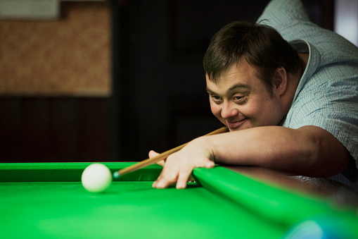 Man with Down syndrome aiming for the white ball in a snooker game, smiling.