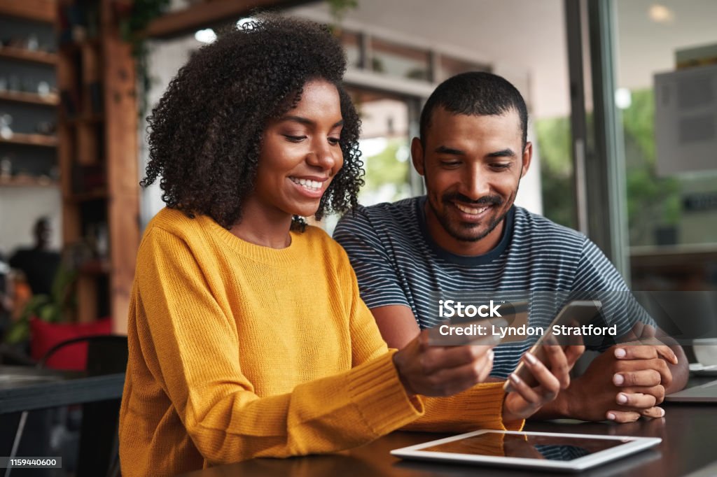 Man looking at his girlfriend shopping online in cafe Young african man looking at his girlfriend using credit card and smartphone for shopping online in cafe Credit Card Stock Photo