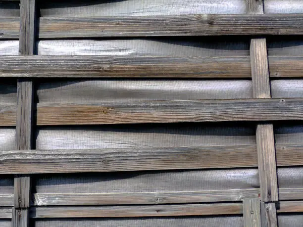thin woven bent wooden slat fence of horizontal strips on vertical frame with vinyl infill. old aged vintage look. weathered texture under bright sunshine.