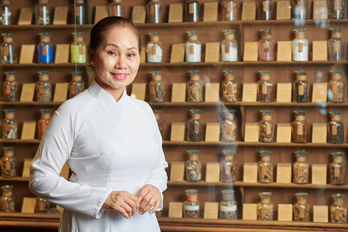 Portrait of positive traditional Asian apothecary owner standing at shevles with glass bottles of herbs, berries and mushrooms