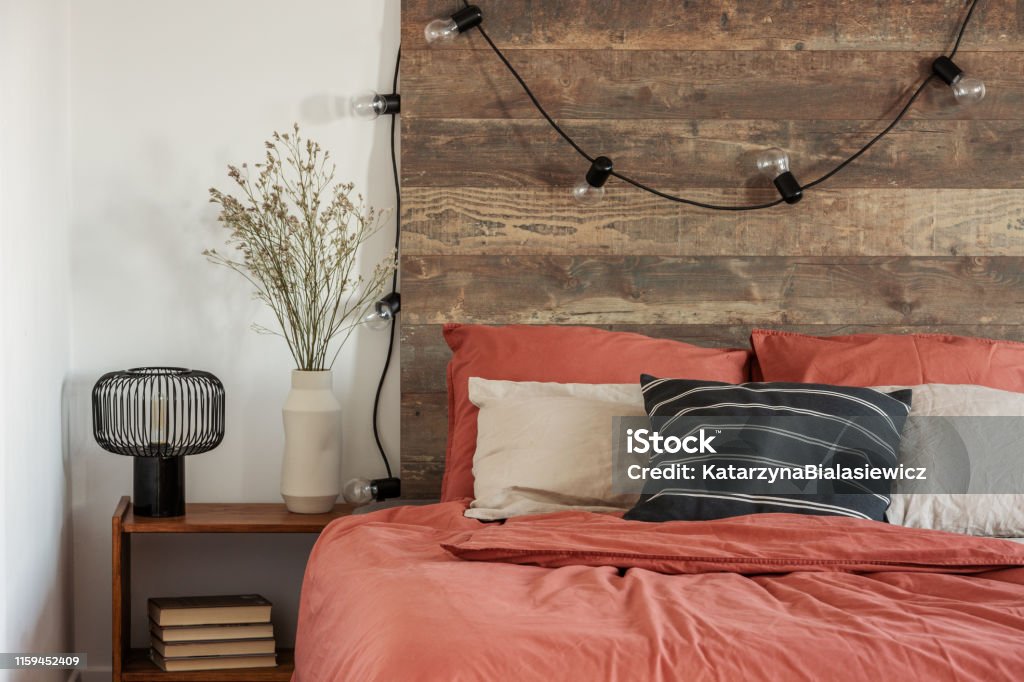 Flowers in beige vase and black industrial lamp on wooden bedside table next to comfortable bed with pillows Apartment Stock Photo