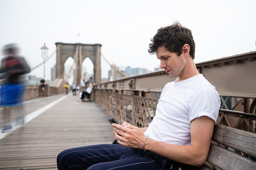 This is a color photograph of a young American man sitting with a phone in hand in New York City, USA.