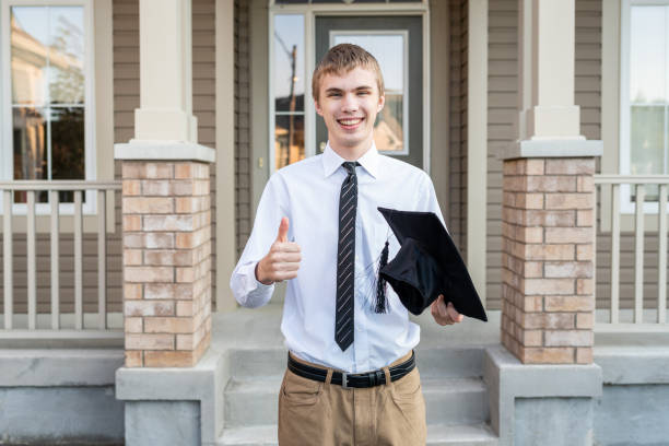 Young male student holding a diploma and a graduation cap in front of a house. stock photo
