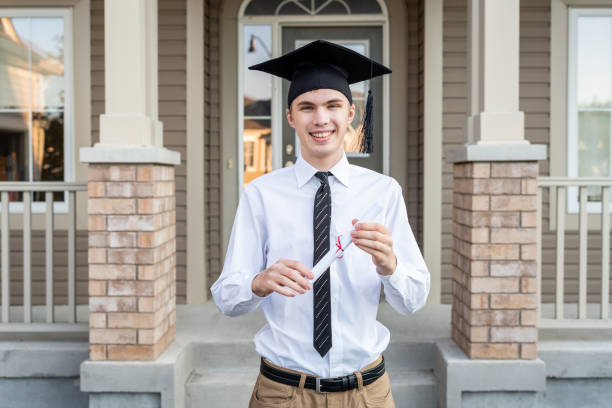Young male student holding a diploma while wearing a graduation cap in front of a house. stock photo