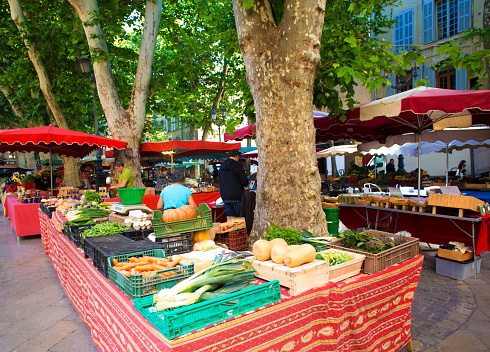 Aix-en-Provence, France: A wide overview of the pretty farmers market on Place Richelme in Aix-en-Provence in early morning in summer. Leafy plane trees tower over the tables, vendors, and red market umbrellas.