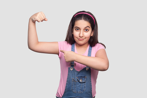 Portrait of satisfied strong brunette young girl in casua pink t-shirt and blue overalls standing looking at camera and pointing at her biceps. indoor studio shot, isolated on light gray background.