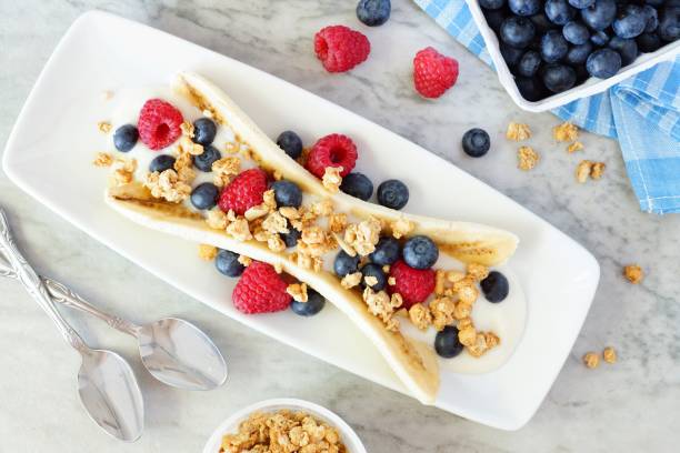 Healthy banana split with yogurt, berries and granola, overhead on marble