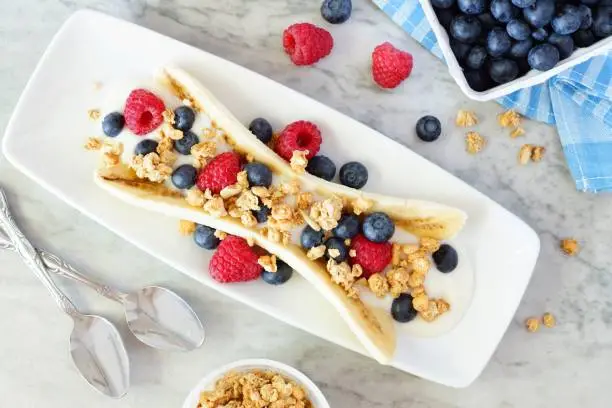 Photo of Healthy banana split with yogurt, berries and granola, overhead on marble