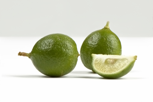 Three Key Limes on a white background.  One is sliced in the foreground.