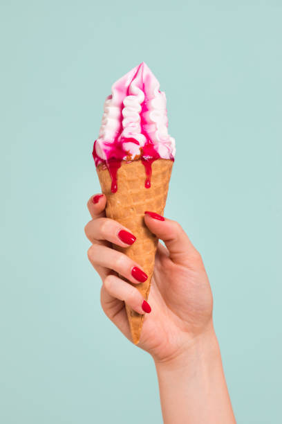 Female hand holds an ice cream cone with cherry jam in a waffle glass on a pink background. Ice cream melted and flowed over fingers and hand
