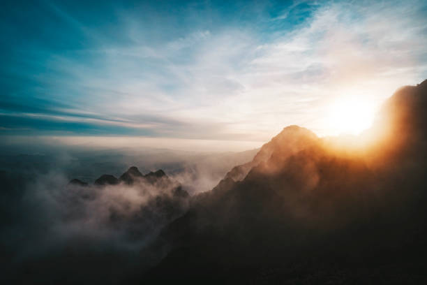 山々からの素晴らしい日の出の眺め。太陽が山の後ろに沈む。高い山の頂上から雲に覆われた谷までの眺め。明るい日差しの中、霧の中で山頂のシルエット。 - 山 ストックフォトと画像