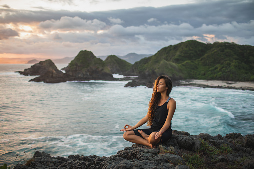 Mujer joven practicando yoga en pose de loto al atardecer con hermosa vista al mar y a la montaña. Sensibilidad a la naturaleza. Autoanálisis y búsqueda del alma. Concepto espiritual y emocional. photo