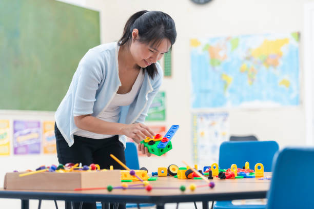 Asian Teacher Cleaning Up Activity A young teacher of Asian descent cleans up the mess after her students did an activity. She is smiling and enjoying looking at their creations. She is casually dressed. teacher classroom child education stock pictures, royalty-free photos & images