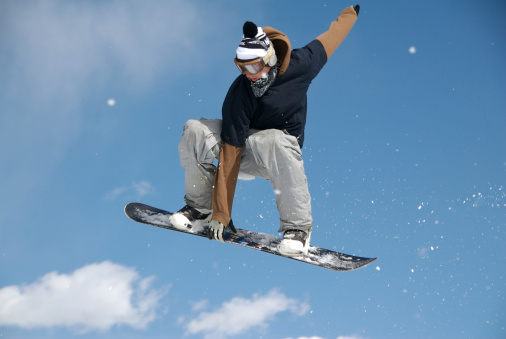 Male Snowboarder with a hat, goggles and a handkerchief   jumping high in the air and grabbing the front edge of his board with his hand with blue sky and a cloud behind him