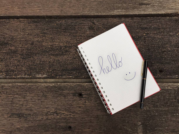 istock Hello text on notebook. Blank open notepad and black pen on old wood table background with copy space. Top view of open note book on grunge wood for business. Word Hello in notebook. 1159512690