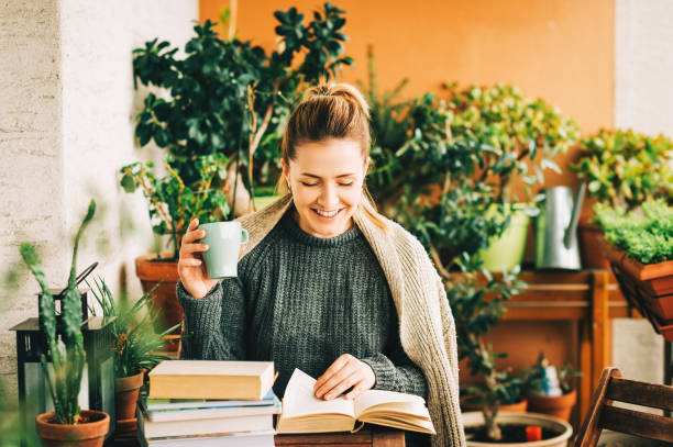 istock Young beautiful woman relaxing on cozy balcony, reading a book, wearing warm knitted pullover, holding cup of tea or coffee 1159513990