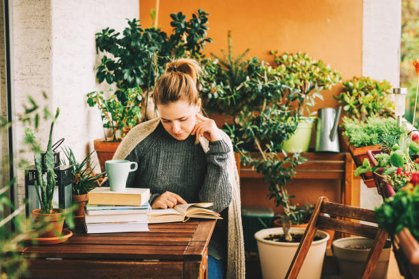 istock Young beautiful woman relaxing on cozy balcony, reading a book, wearing warm knitted pullover, cup of tea or coffee on stack of books 1159514009