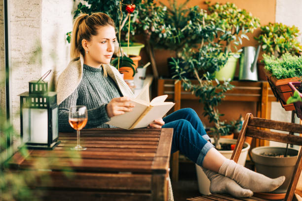 istock Young beautiful woman relaxing on cozy balcony, reading a book, wearing warm knitted pullover, glass of wine on wooden table 1159514013