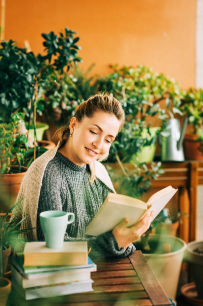 istock Young beautiful woman relaxing on cozy balcony, reading a book, wearing warm knitted pullover, cup of tea or coffee on stack of books 1159514026