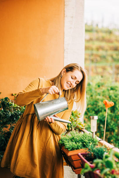 Portrait of beautiful woman watering green plants on the balcony, wearing brown cotton dress stock photo
