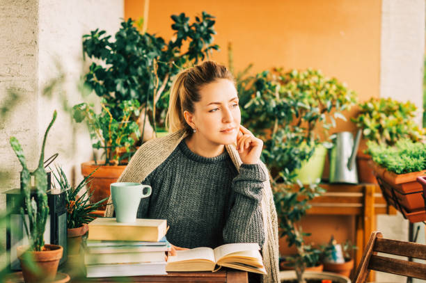 istock Young beautiful woman relaxing on cozy balcony, reading a book, wearing warm knitted pullover, cup of tea or coffee on stack of books 1159514049