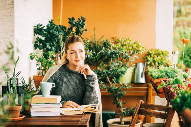 istock Young beautiful woman relaxing on cozy balcony, reading a book, wearing warm knitted pullover, cup of tea or coffee on stack of books 1159514050