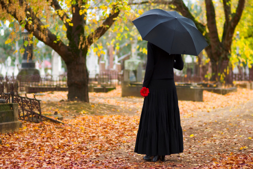 A lone woman in mourning in a cemetery in fall (autumn), dressed in black and carrying a black umbrella and a red flower. Surrounding deciduous trees give the image a yellow and orange background and enhance the sense of loss and sadness.