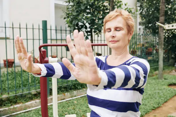 Aged woman outstretching arms in front of her, closing eyes and doing breathing exercise