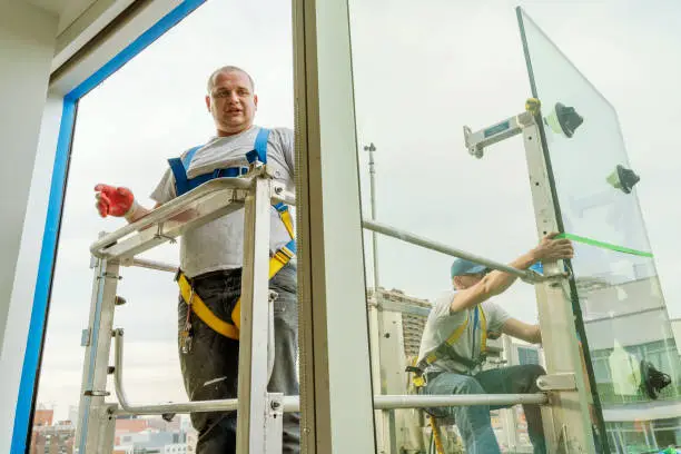 Team of blue-collar workers replacing a broken window in the office building. High-altitude work on the lifting platform which is placed outside.