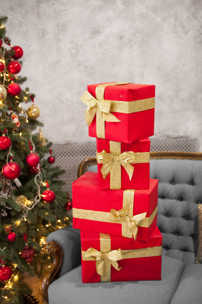 istock Red boxes with gold ribbons with bows standing on each other on couch with christmas tree with red decorations in the room. Christmas postcard concept. Blog content. 1159532297