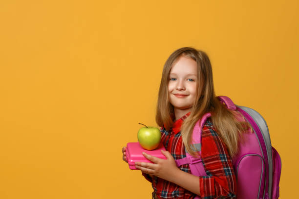 portrait of a little girl schoolgirl on a yellow background. the child has a satchel behind his back and holds a lunchbox in his hands. back to school. the concept of education. - lunch box lunch red apple imagens e fotografias de stock