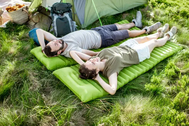 Young and cheerful couple lying on the mattress at the campsite, enjoying summer time while traveling in the mountains