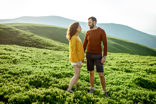 Lovely couple in bright sweaters standing together on the green meadow, while traveling high in the mountains during the sunset